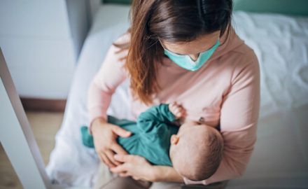 Mother wearing a surgical mask while nursing her newborn. Caring for Babies and Small Children During COVID-19.