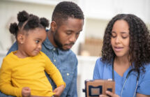 A female doctor of African descent is showing a father and toddler daughter the Children's Medical Records. The child is sitting on her father's lap. The doctor is holding a tablet computer.