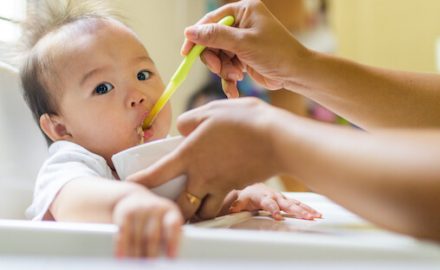 Young Asian baby sitting in a highchair with adult spoon feeding. Babies start solids at different ages, depending on their development.