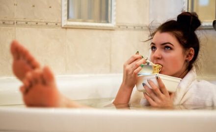 Young woman wearing a bathrobe while sitting in a bathtub eating noodles. What we eat during pregnancy changes food preferences.
